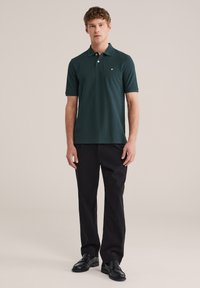 Jonge man staand, gekleed in een donkergroen poloshirt met korte mouwen, zwarte broek en zwarte loafers, tegen een effen beige achtergrond.