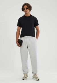 Czarny T-shirt z krótkim rękawem, szare dresowe spodnie z troczkami, beżowe sneakersy. Model trzyma małą czarną saszetkę. Gładkie tło.