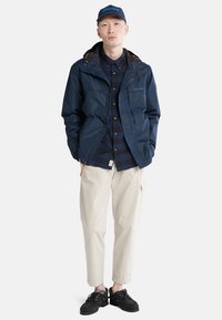 Giacca impermeabile blu navy con dettagli arancioni, due tasche frontali, camicia a quadri nera, pantaloni beige e sneakers nere.
