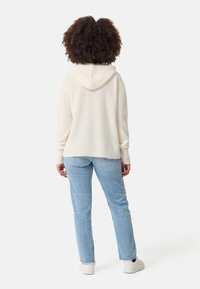 Cremfarbener Kapuzenpullover mit strukturiertem Strickdesign, kombiniert mit hellblauen Jeans. Weiße Sneakers runden den lässigen Look ab.