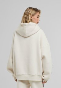 Felpa oversize color crema in morbido tessuto con cappuccio regolabile, dotata di polsini e vita a costine. Senza loghi o decorazioni visibili.