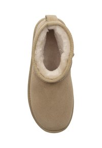 Beżowy kapcie z zamszowym wykończeniem, miękką białą podszewką z owczej wełny, okrągłym kształtem i niskim fasonem sięgającym kostki. Posiadają przeszyty brzeg i boczną pętlę.