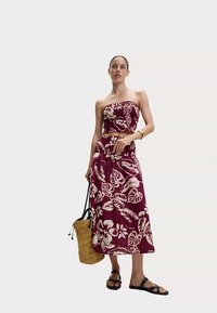 Conjunto burdeos de dos piezas con estampado floral; top corto sin tirantes y falda larga. Los accesorios incluyen un bolso tejido y sandalias negras.