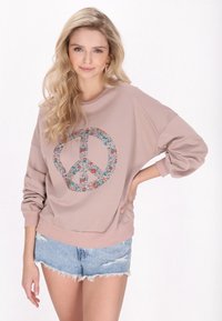 Felpa a maniche lunghe di colore rosa chiaro con un design di simbolo di pace floreale. Presenta una vestibilità ampia e polsini a coste, abbinata a pantaloni corti in denim strappati.