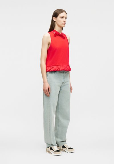 Γυναίκα όρθια, φοράει αμάνικη κόκκινη μπλούζα με φιόγκο και βολάν στη βάση, ανοιχτό μπλε τζιν κανονικής γραμμής και πολύχρωμα αθλητικά παπούτσια σε λευκό φόντο.