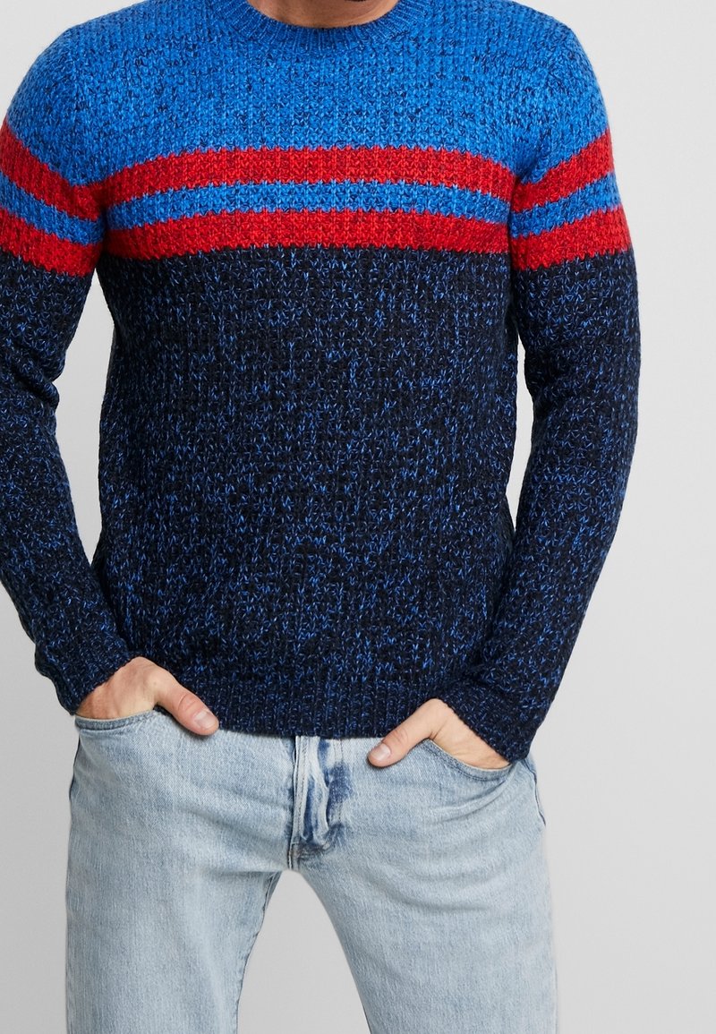 Sweter z dzianiny z niebieską górną częścią, czerwonymi poziomymi paskami i ciemnoniebieską dolną częścią. Wykonany z teksturowanej włóczki, z okrągłym dekoltem.