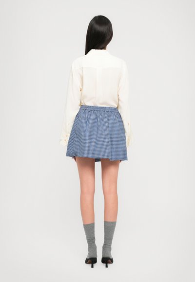 Blusa blanca con botones combinada con una falda a cuadros azul. Medias grises hasta la rodilla y zapatos de tacón negros completan el conjunto.