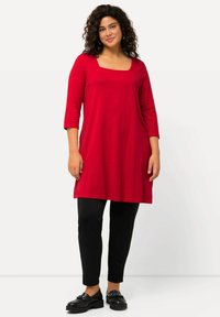 Rote langärmlige Tunika mit einem quadratischen Ausschnitt, kombiniert mit schwarzen engen Hosen. Der Stoff wirkt weich mit einer glatten Textur, akzentuiert durch schwarze Schuhe.