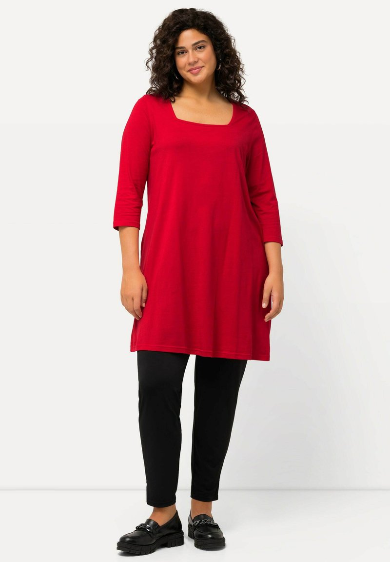 Rote langärmlige Tunika mit einem quadratischen Ausschnitt, kombiniert mit schwarzen engen Hosen. Der Stoff wirkt weich mit einer glatten Textur, akzentuiert durch schwarze Schuhe.