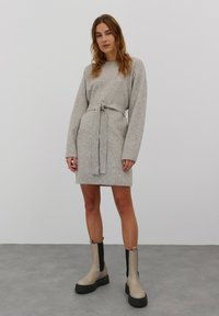 Hellgraues Strickkleid mit langen Ärmeln und einem Gürtel, versehen mit gerippten Akzenten an den Bündchen und dem Saum. Kombiniert mit beigefarbenen und schwarzen Ankle-Boots.