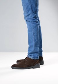 Stivaletti alla caviglia in camoscio marrone con suola in gomma nera, abbinati a jeans denim blu aderenti, che mostrano una texture liscia e un design pulito.