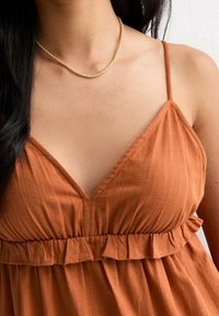 Robe couleur rouille avec un décolleté en V et une taille volants. Fabriquée en tissu texturé, avec de fines bretelles et une coupe décontractée. Collier en or visible.