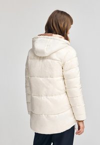 Chaqueta tipo puff en crema con capucha, que presenta secciones acolchadas horizontales, una textura suave y un forro interior en contraste.