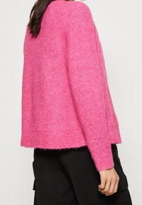 Luddig rosa tröja med en något lös passform, ribbat halslinne och sydd fåll. Matcher med svarta tygbyxor med fickor.