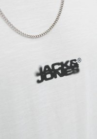 White cotton t-shirt featuring a black, faded graphic logo "JACK & JONES" on the front. Accompanied by a silver chain necklace.