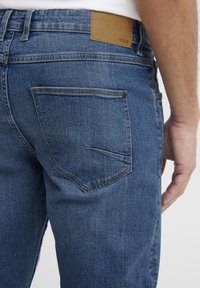 Jeans de mezclilla azul con una textura suave, que cuentan con una etiqueta de cuero marrón que dice "ISOLID" y un bolsillo trasero con un detalle de costura curvada.
