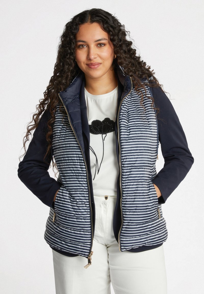 Marineblauer und gestreifter Steppweste mit Reißverschluss, Seitentaschen und taillierter Passform; kombiniert mit einem weißen Shirt mit floralem Grafikdesign.