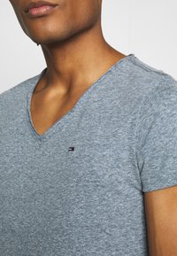 Grå t-shirt med korta ärmar och v-ringning i ett mjukt, texturerat tyg. Har en liten, f ärgglad logga på vänster bröstområde.