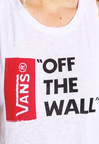 Bílý tílko s červeným obdélníkovým logem s textem "VANS" a frází "OFF THE WALL" tučným černým písmem. Měkká textura látky.