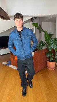 Mladý muž v modré džínové bundě, černých kalhotách a černých botách stojí v obývacím pokoji s velkou rostlinou a oranžovou pohovkou v pozadí.