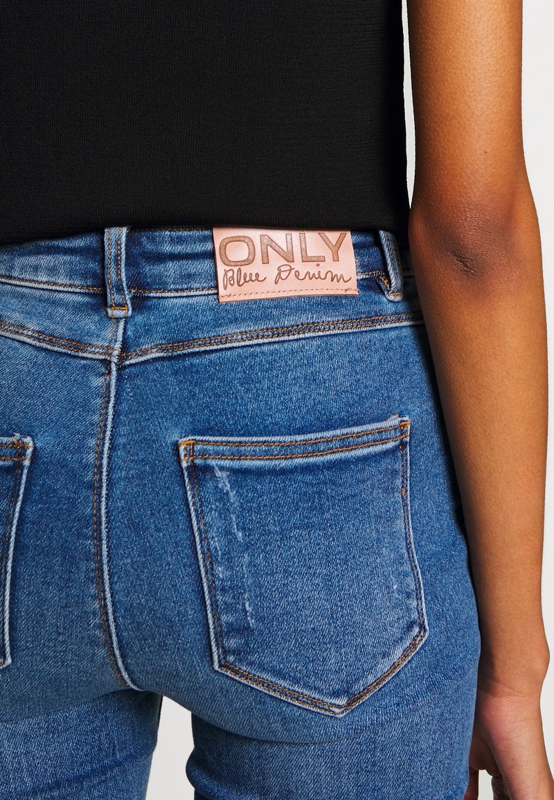 Personne portant un jean en denim bleu avec un patch en cuir beige inscrit "ONLY Blue Denim" sur la ceinture arrière et un haut noir.