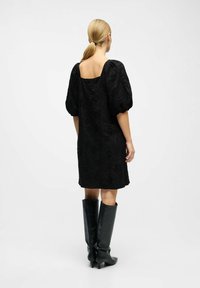 Vestido negro con mangas abullonadas, escote cuadrado en la espalda, patrón floral texturizado, hasta la rodilla, combinado con botas de cuero negro hasta la rodilla.