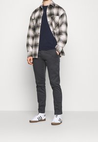 Rutig flanellskjorta i beige och svart, marinblå t-shirt, mörkgrå byxor, vita sneakers med svarta ränder. Modell poserar avslappnat.