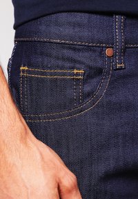 Mörkblå denimjeans med gyllene gula sömmar. Har en framficka och en bronsnitar, vilket visar på en klassisk passform och hållbart material.