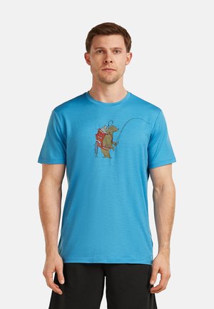 Mann trägt ein blaues T-Shirt mit einer Grafik eines fischenden Bären, einen Rucksack und einen Hut, steht vor weißem Hintergrund.