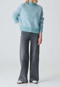 Personne portant un pull bleu clair en laine pelucheuse, un jean noir délavé à jambes larges et des chaussures blanches, debout devant un fond uni.