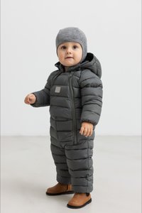 Grå puffer snødrakt med hette, med quiltede seksjoner, frontglidelås og et logo-patch. Brukt med en grå strikket lue og brune støvler.