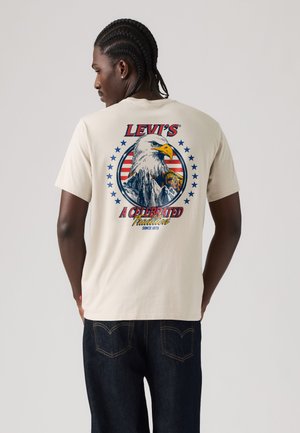 Homme aux cheveux tressés portant un t-shirt beige Levi's avec un motif aigle, montagne et étoiles, et un jean bleu foncé, tourné de dos à la caméra.