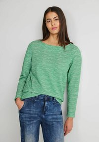 Grünes Langarmshirt mit strukturiertem, gestreiftem Muster; runder Ausschnitt; getragen mit Jeans aus Denim mit leichter Ausbleichung und Taschen.