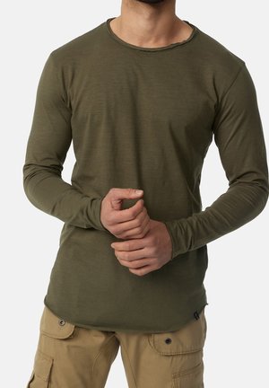 Homme portant une chemise à manches longues vert olive et un pantalon cargo beige, mains jointes à la taille, torse visible sur un fond uni.
