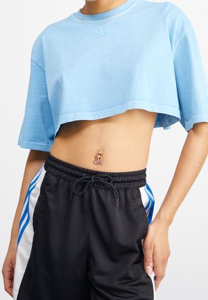 Osoba nosząca jasnoniebieski krótki t-shirt i czarne sportowe szorty z niebieskimi i białymi paskami, pokazująca piercing w pępku.