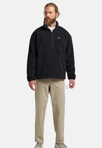 Pull-over en polaire noire avec col zippé, au design simple. Associé à un pantalon beige et des baskets de couleur claire.