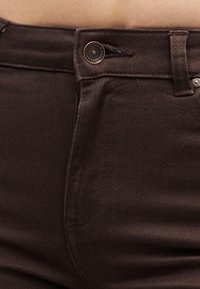 Gros plan sur une personne portant un jean marron foncé, centré sur la ceinture, le bouton, la fermeture éclair et les passants de ceinture.
