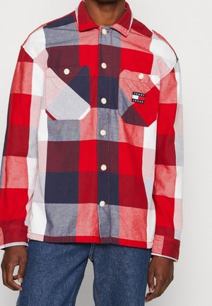 Mężczyzna ubrany w czerwoną, białą i granatową kratkę na koszuli zapinanej na guziki z dwoma zapinanymi kieszeniami na piersi, z których jedna ma metkę "Tommy Jeans", w połączeniu z niebieskimi dżinsami.