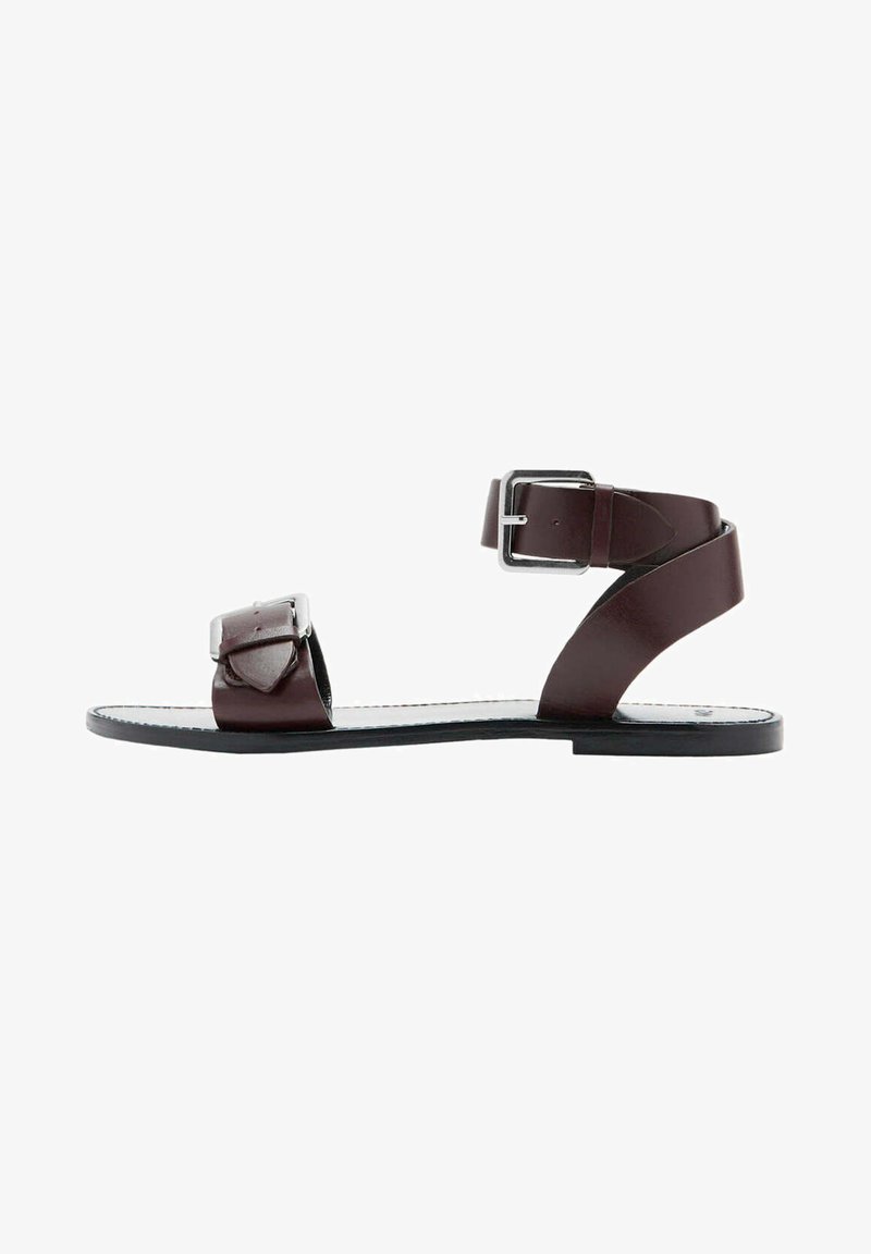 Sandale plate en cuir marron avec deux boucles argentées réglables, une au-dessus des orteils et une autour de la cheville, sur une semelle noire avec une couture blanche.