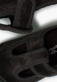 Zwarte textielschoenen met klittenbandsluitingen, voorzien van uitsparingen aan de zijkant en een zachte, gestructureerde afwerking. Duurzame stiksels zijn zichtbaar als accenten.