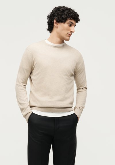 Άνδρας με σγουρά σκούρα μαλλιά, που φοράει μπεζ πουλόβερ πάνω από λευκό πουκάμισο και μαύρο παντελόνι, στέκεται με τα χέρια στις τσέπες, κοιτάζοντας προς το πλάι.