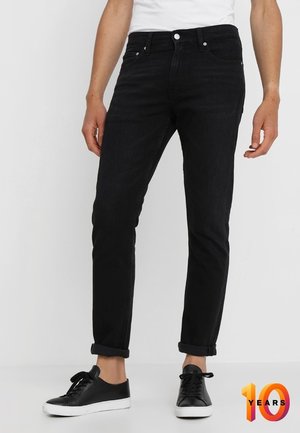Personne portant un jean slim noir retroussé et des baskets noires avec des semelles blanches, debout devant un fond clair uni.