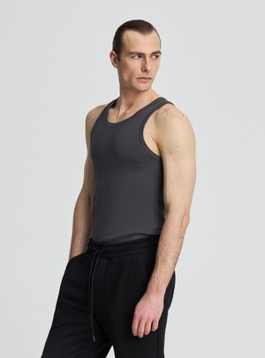 Uomo con capelli pettinati all'indietro che indossa una canotta grigio scuro senza maniche e pantaloni neri con cordino, guarda verso sinistra su uno sfondo neutro.