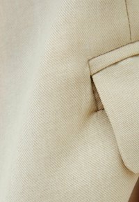 Béžová tkanina s texturovaným vazem, s přehnutým detailem podél okraje. Materiál se zdá být hladký s viditelným švem.