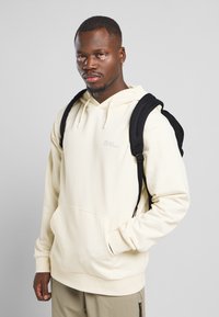 Sudadera con capucha color crema, con un bolsillo frontal, hecha de una tela suave, llevada sobre pantalones verdes, acompañada de una mochila negra.