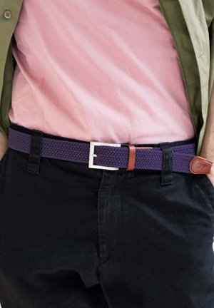 Uomo che indossa pantaloni neri, una cintura viola intrecciata con dettagli in pelle e una camicia rosa infilata sotto una giacca verde.