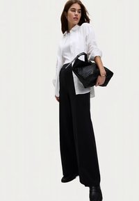 Hvid skjorte med knapper, sorte højtaljede bukser med vide ben, sort flettet håndtaske og sorte sko. Modellen står foran en neutral baggrund.