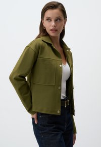 Veste vert olive en tissu lisse, dotée d'un col, de boutons à pression et d'une poche poitrine. Associée à un top blanc et à un jean foncé.