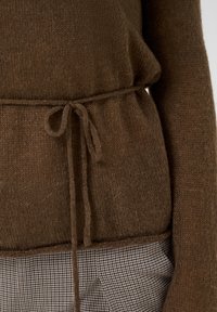 Pull en tricot marron à manches longues noué à la taille, associé à un pantalon marron et blanc à petits carreaux.