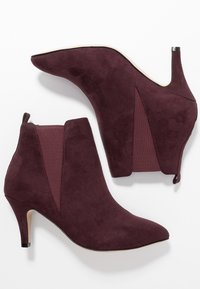 Stivaletti alla caviglia in suede bordeaux con punte appuntite, pannelli laterali elasticizzati e tacchi da 3 pollici. Presentano una superficie liscia e una silhouette elegante.